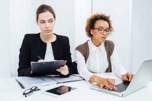 Two focused young business women working using clipboard and laptop